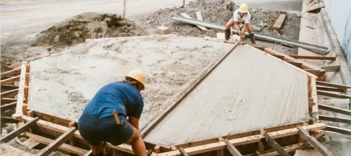 Dva dělníci hladí čerstvý beton v dřevěných tvářnicích základové desky.