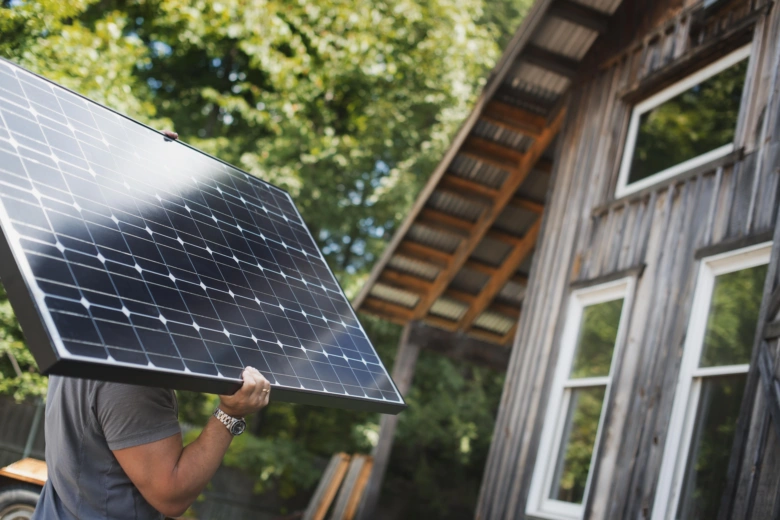 Muž nese fotovoltaický panel k dřevěnému domu.