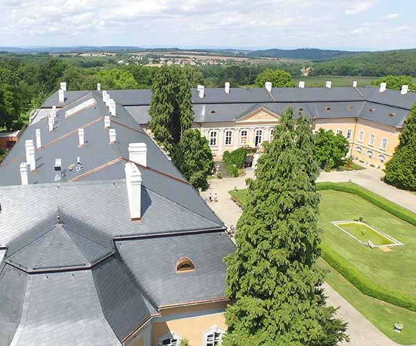 Velká historická budova s břidlicovou střechou na pozadí zelených kopin.