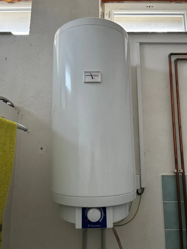 Bílý elektrický bojler namontovaný na zdi s viditelnými potrubím.