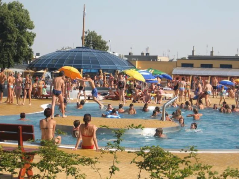 Lidé plavou v bazénu na koupališti s barevnými slunečníky.