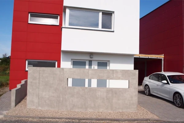Pohledový beton na plot u moderního domu s červenou fasádou.