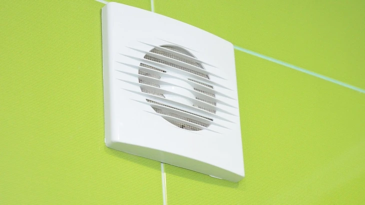 Bílý ventilátor pro odvětrání koupelny namontovaný na zelené dlažbě.