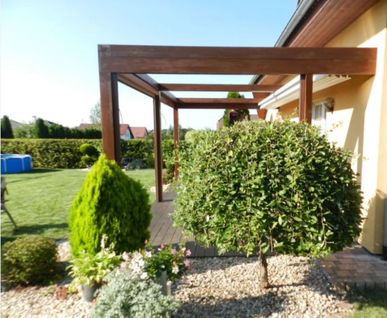 Moderní dřevěná pergola u domu s okrasnými rostlinami v zahradě.