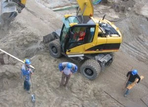 Žlutý bagr pracuje na staveništi s třemi dělníky v modrých uniformách.