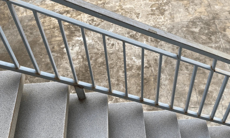 Světle šedé kovové zábradlí na betonovém schodišti s dlažbou.