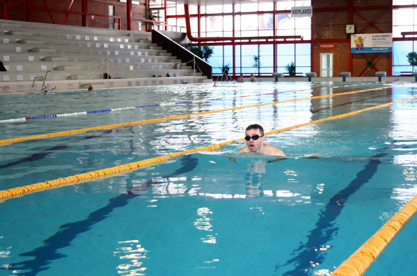 Muž plave na zádech v krytém bazénu s žlutými čarami a tribunami.