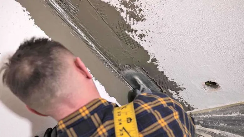 Muž lepi perlinku na roh stěny špachtlí s hmotou.