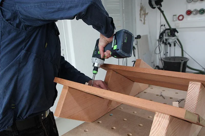 Muž montuje dřevěné schody v dílně pomocí elektrického šroubováku.
