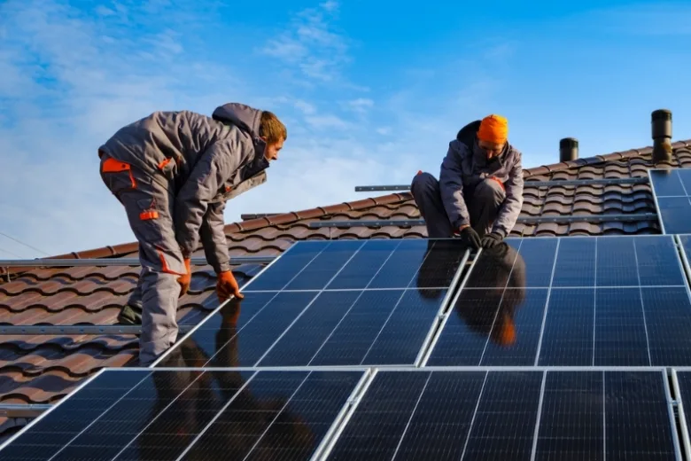 Dva dělníci montují fotovoltaické panely na střechu domu.