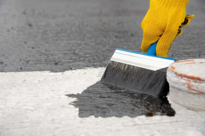 Ruka v žluté rukavici nanáší tekutou hydroizolaci štětcem na beton.