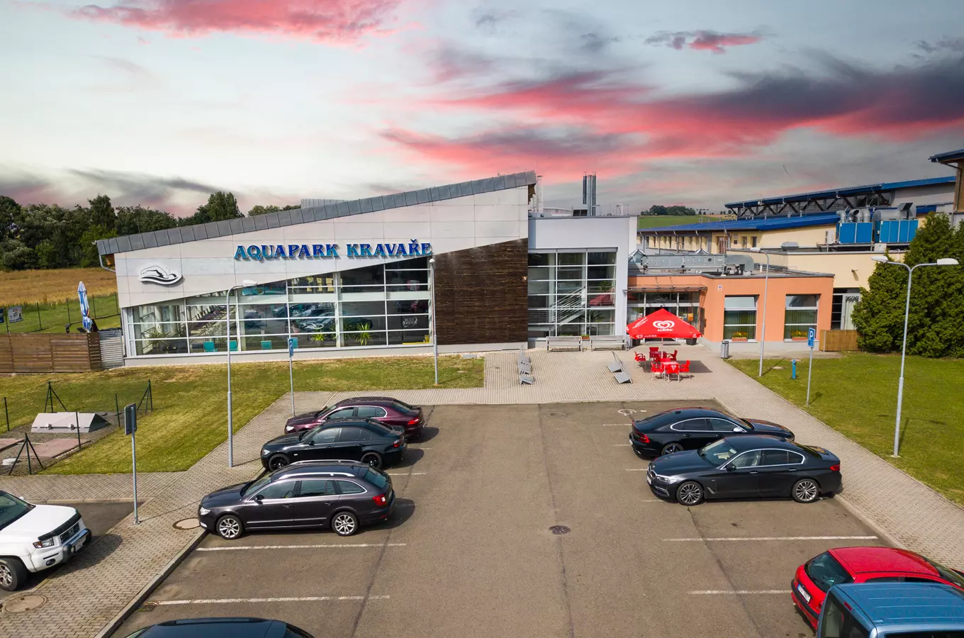 Vnější pohled na Aquapark Kravaře s parkovištěm aut pod západním sluncem.