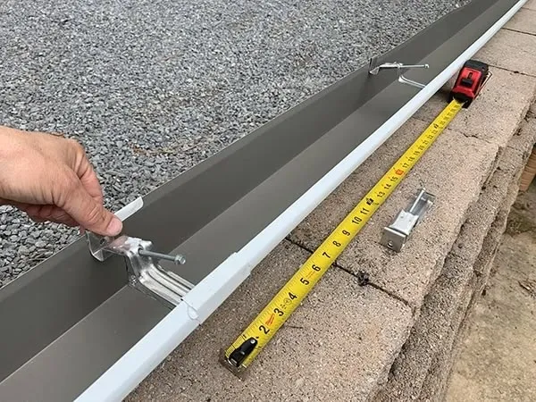 Ruční montáž okapového háku na bílý spádový kanál s měřicí páskou.
