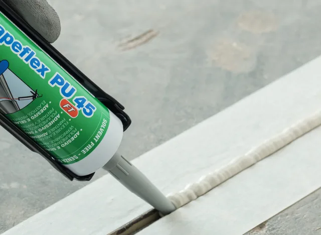 Ruka aplikuje silikonový tmel do spáry na bílém povrchu.