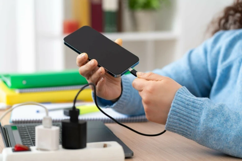 Osoba nabíjí chytrý telefon pomocí kabelu na pracovním stole.