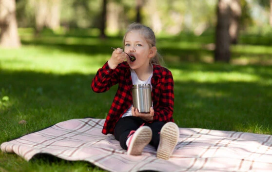 Dívka sedí na plátně v parcí a jí z termosky s mrazeným dezertem.