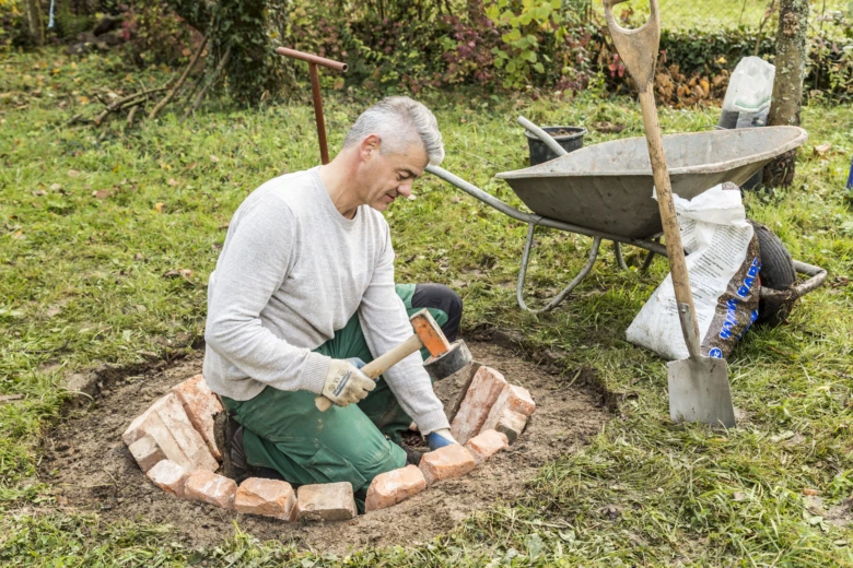 Muž staví ohniště z cihel svépomocí v zahradě.