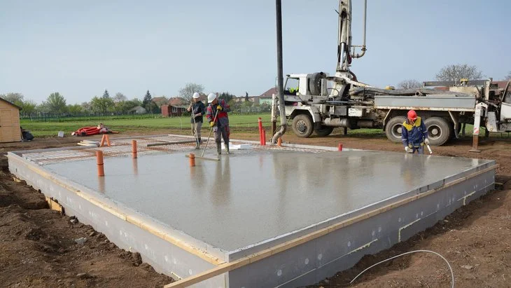 Práce na betonové základní desce s pracovníky a stavebním vozem.