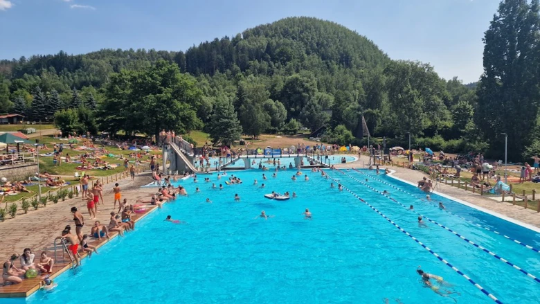 Venkovní bazén Košťálov s plavci a diváky na sluneční louce.