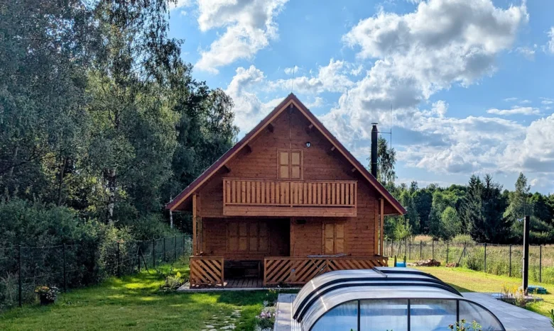 Dřevěná chata s terasou a skleníkem na pozadí zelené zahrady.