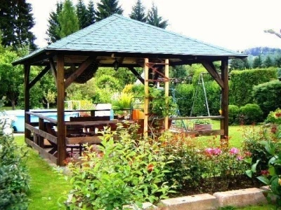 Dřevěná zahradní pergola s posezením uprostřed zelené zahrady.