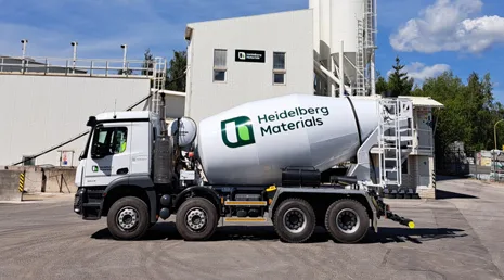 Bílý betonový míchač značky Heidelberg Materials před továrnou.