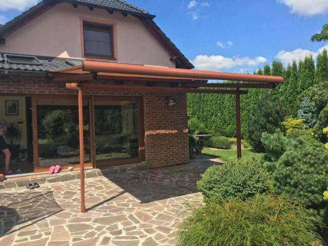 Dřevěná pergola stínící kamennou terasu u domu.